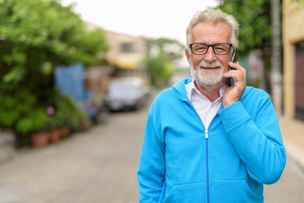 Happy handsome senior bearded man smiling while talking on mobile phone and wearing eyeglasses outdoors